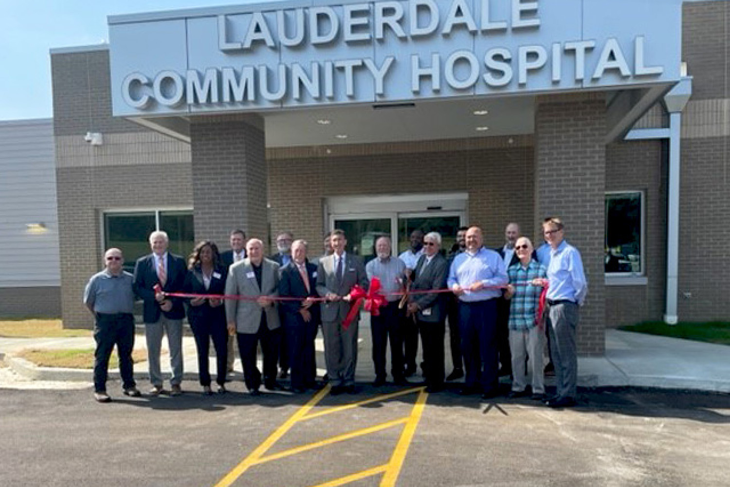 A group of people stand in front of Lauderdale Community Hospital, holding a red ribbon at a ribbon-cutting ceremony. The hospital's name is displayed above the entrance, and the event appears to be taking place during the day.