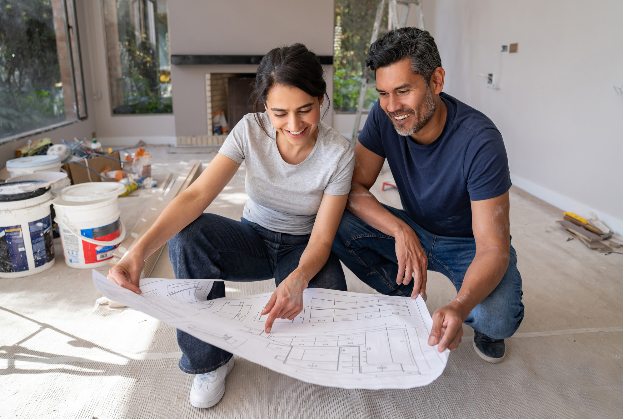 Un hombre y una mujer sonrientes se arrodillan en el suelo de una habitación parcialmente renovada, discutiendo planos, rodeados de cubos y herramientas, mientras planifican su proyecto, posiblemente considerando la posibilidad de solicitar un préstamo con garantía hipotecaria para financiar su renovación.