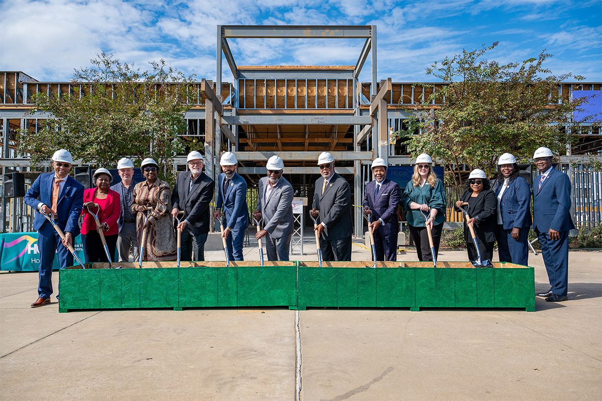 Un grupo de personas con cascos de protección se encuentran delante de una obra en construcción, cada una con una pala en la mano, preparadas para romper el suelo. Están vestidos de etiqueta y alineados en una zona pavimentada bajo un cielo parcialmente nublado.