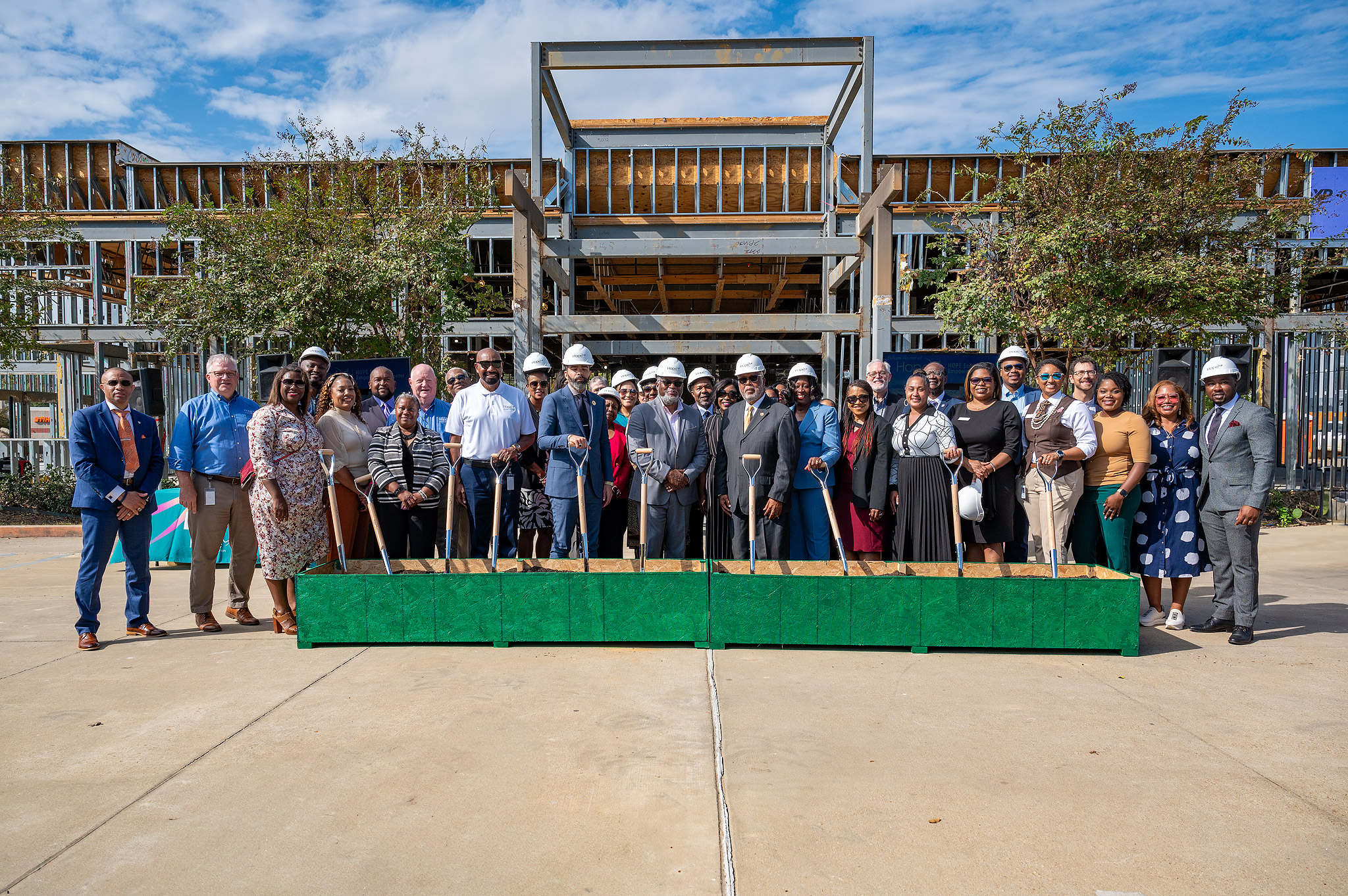 Un grupo de personas vestidas de etiqueta y con cascos permanecen de pie delante de un edificio en construcción. Llevan palas en las manos, lo que sugiere una ceremonia de colocación de la primera piedra. El fondo muestra un cielo parcialmente nublado y árboles.