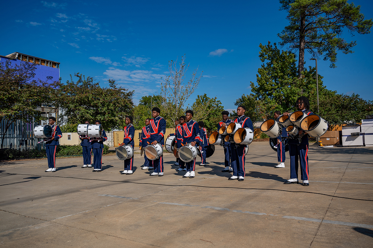 Una banda de música formada por nueve personas vestidas con uniformes azules y rojos actúa al aire libre. Tocan varios tambores en un día soleado, con árboles y un edificio visibles al fondo.