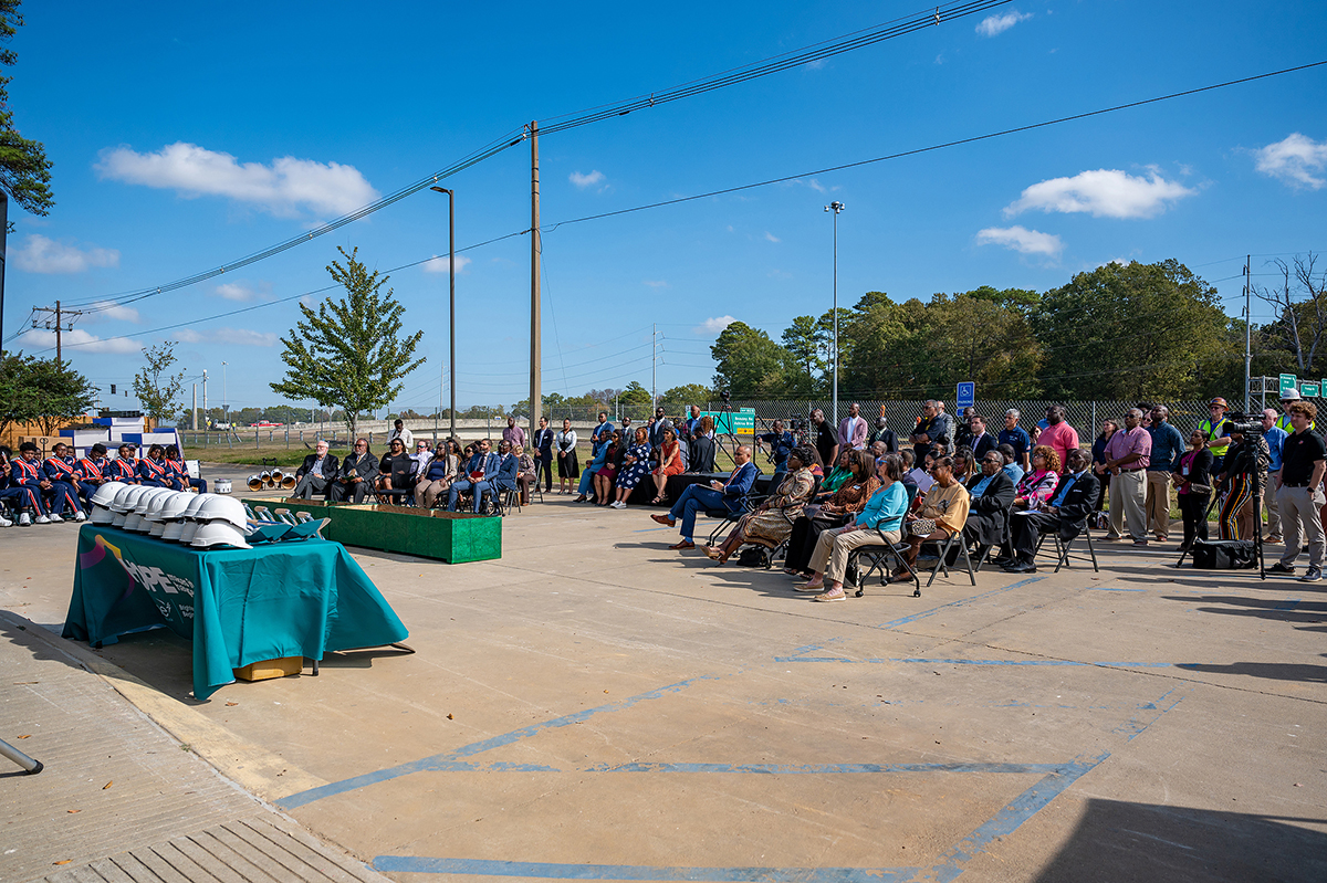 Un grupo de personas sentadas al aire libre en una ceremonia o evento. En primer plano, una mesa con un mantel verde y varios objetos. Al fondo se ven árboles y líneas eléctricas bajo un cielo azul despejado.