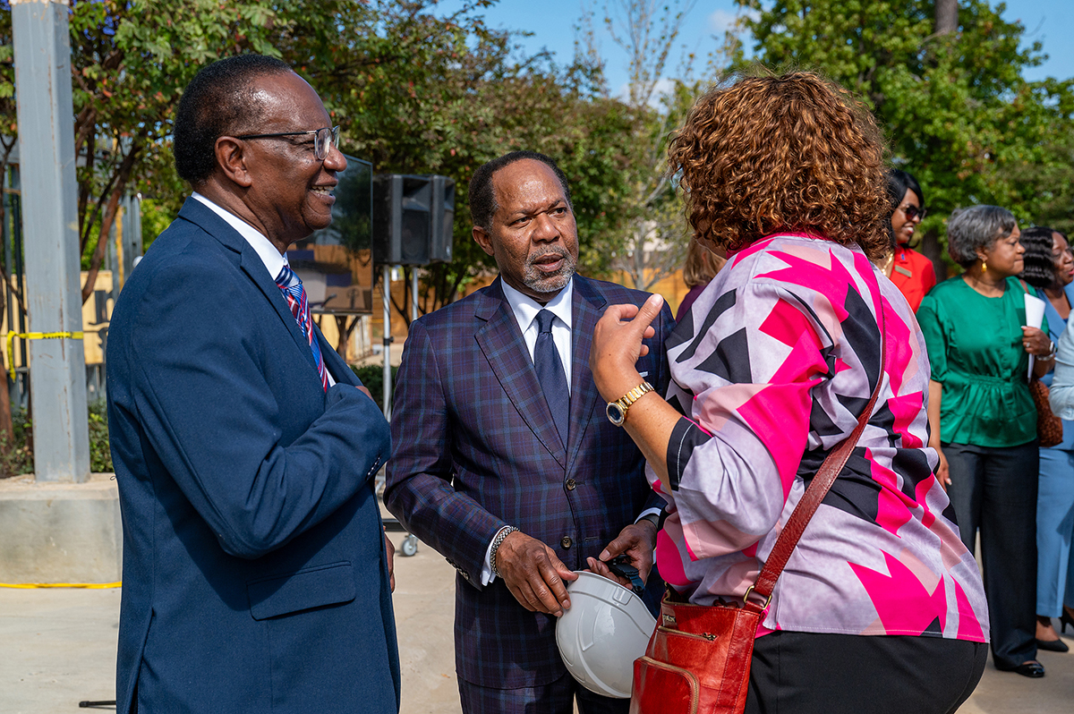 Tres personas conversan al aire libre. Dos hombres trajeados y una mujer con una blusa de colores conversan. Un hombre sostiene un casco. Al fondo se ven otras personas y árboles.