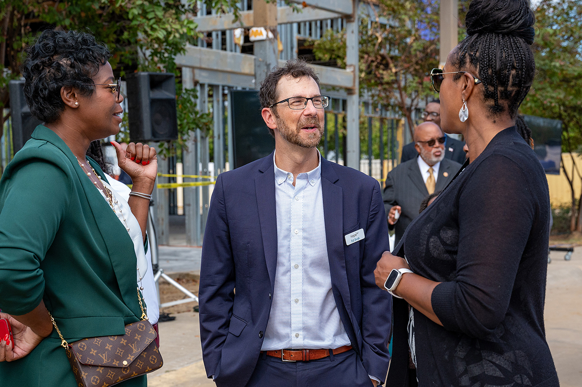 Tres individuos conversan al aire libre: una mujer con americana verde, un hombre con traje azul y una mujer con traje negro. Hay otras personas al fondo, entre ellas una persona con traje y corbata, de pie cerca de la estructura de un edificio.