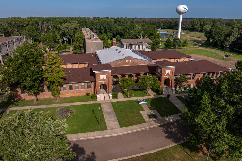 Aerial view of a brick academic building with arched entryways, surrounded by lawns, trees, sidewalks, and nearby campus structures. A white water tower stands in the background against a wooded area.