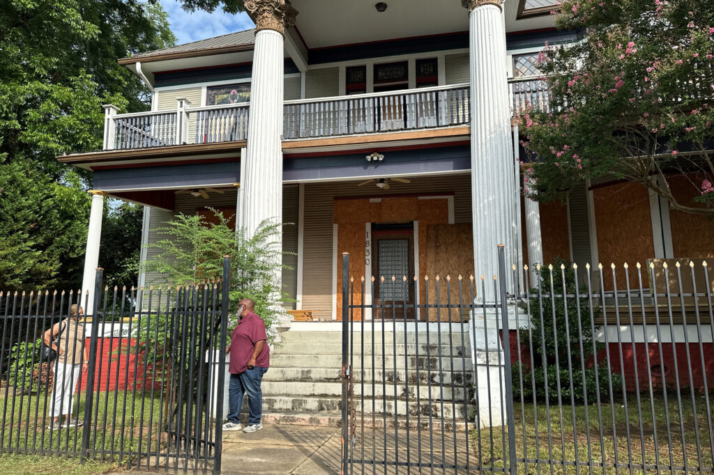 Two people stand outside a large, two-story house with white columns and a boarded-up door and windows. The house is behind a black metal fence, with overgrown plants and trees nearby.