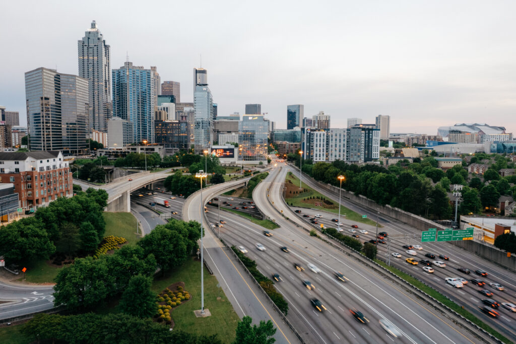 A wide view of a busy highway with multiple lanes and cars, surrounded by green trees and tall modern buildings in a city skyline under an overcast sky.