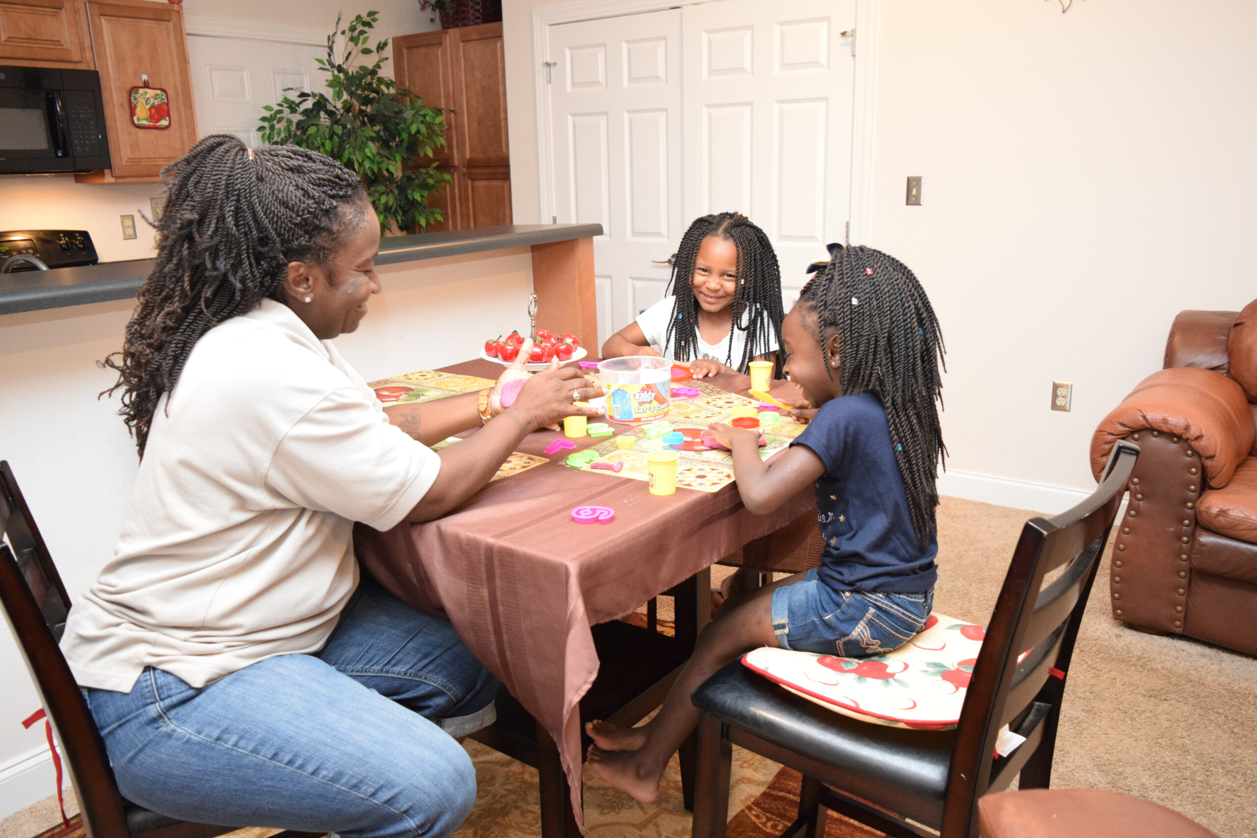 An adult and two children with braided hair sit around a dining table playing with colorful playdough in a cozy kitchen and living room setting. All three appear engaged and smiling.
