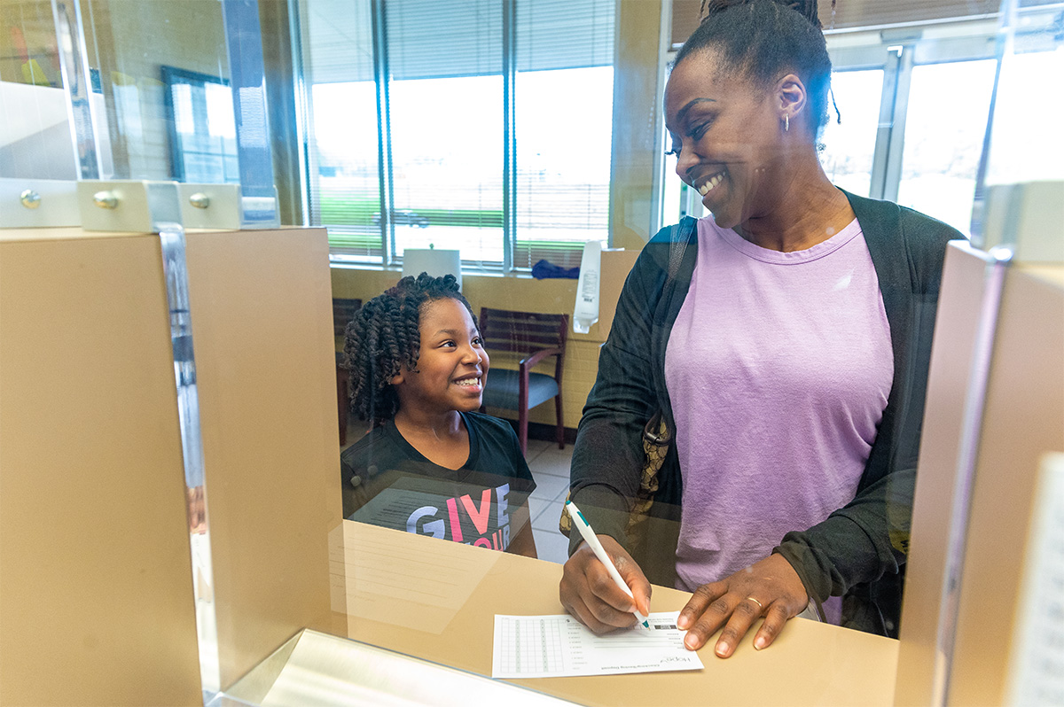 A woman smiles as she fills out a form at a counter while a young girl stands beside her, also smiling. They are separated from the staff by a clear divider in a brightly lit office or reception area.