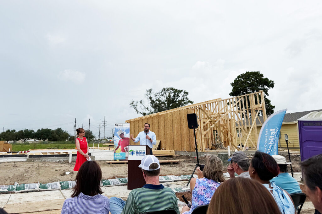 A man speaks at a podium with a Habitat for Humanity sign in front of a partially-built house, as an audience listens. A woman in a red dress stands nearby, and construction materials are scattered around the site.