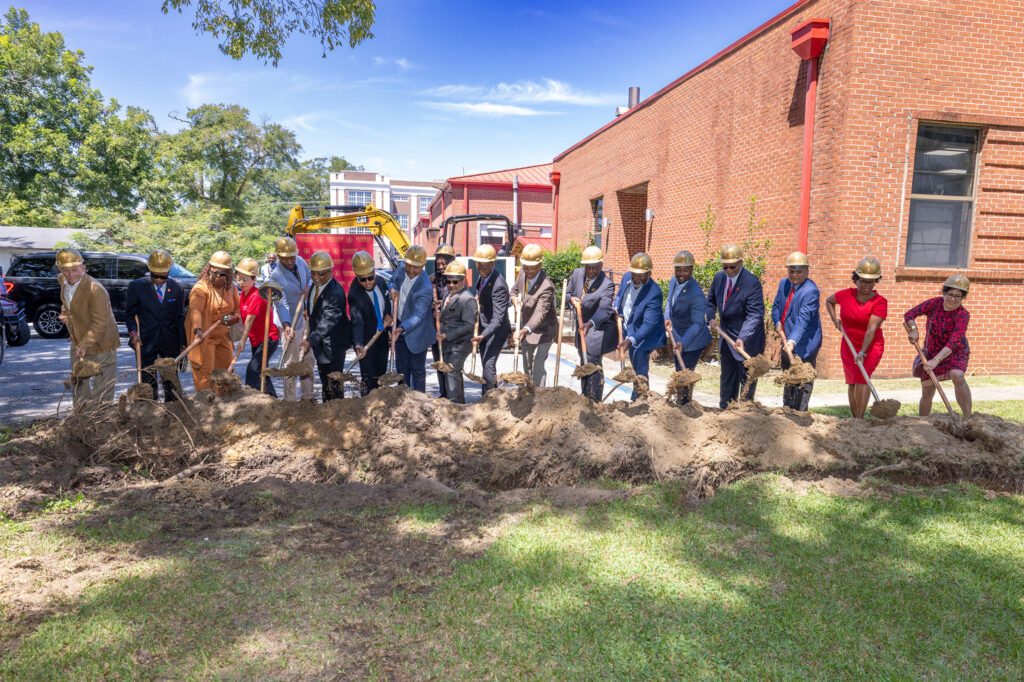A group of people in hard hats and formal attire stand in a line, holding shovels and breaking ground on a dirt patch beside a red brick building, marking the start of construction. Construction equipment is visible in the background.