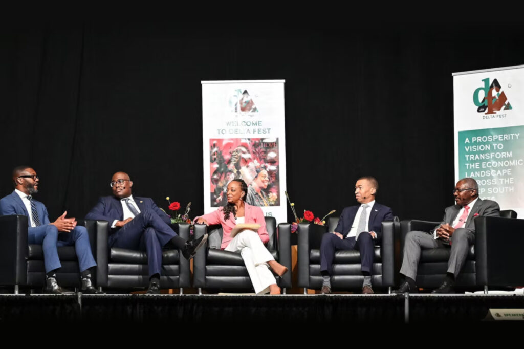 Five people in business attire sit on stage armchairs, engaged in a panel discussion. A woman in pink speaks into a microphone, while four men listen. Event banners with text and images are displayed behind them.