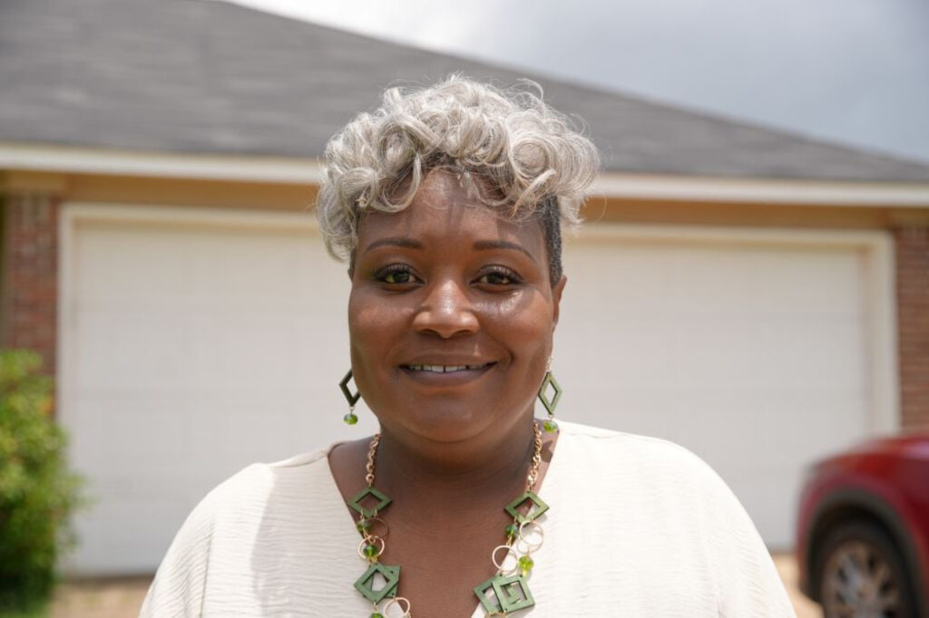 A woman with short, curly gray hair smiles at the camera. She is wearing a white top and green geometric earrings and necklace, standing outdoors in front of a garage door and a red car.