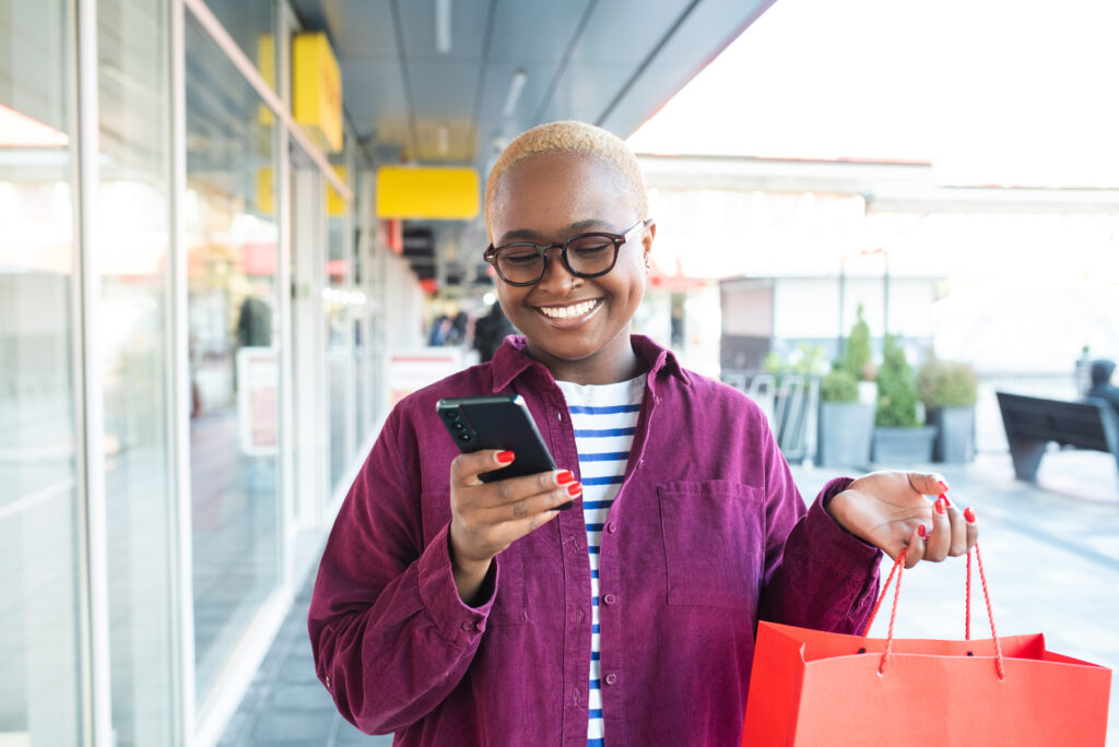 A smiling person wearing glasses and a maroon shirt holds a red shopping bag in one hand and looks at their smartphone with the other while standing outdoors near shop windows.