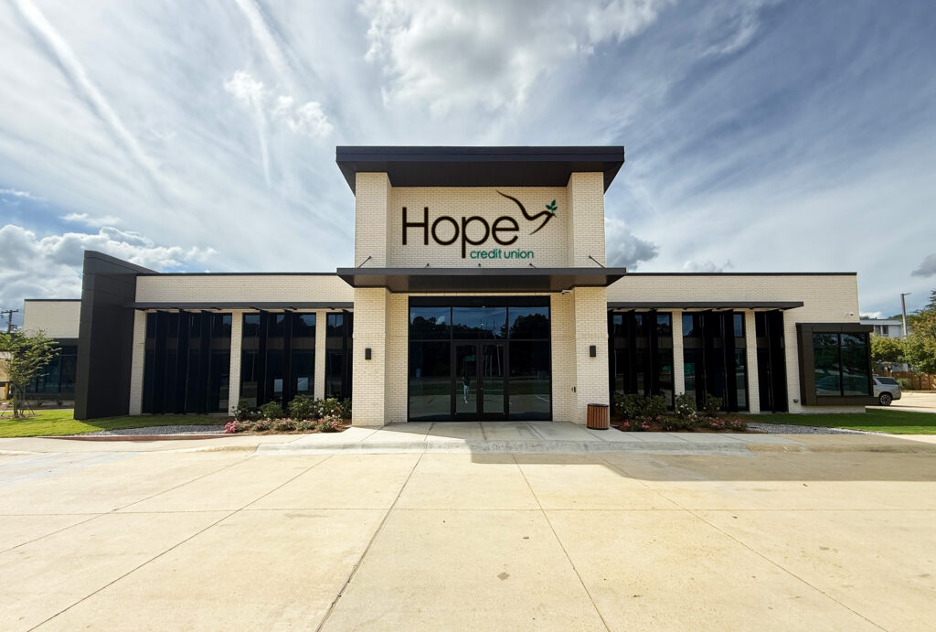 Modern, single-story building with large windows and "Hope Credit Union" sign above the entrance, set against a partly cloudy sky with landscaped greenery and an empty parking lot in front.