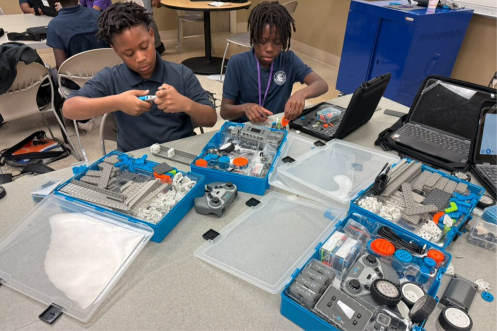 Two children sit at a table assembling robotics kits with various parts and tools. Open boxes of components and laptops are on the table, and both kids are focused on building. Other students are visible in the background.