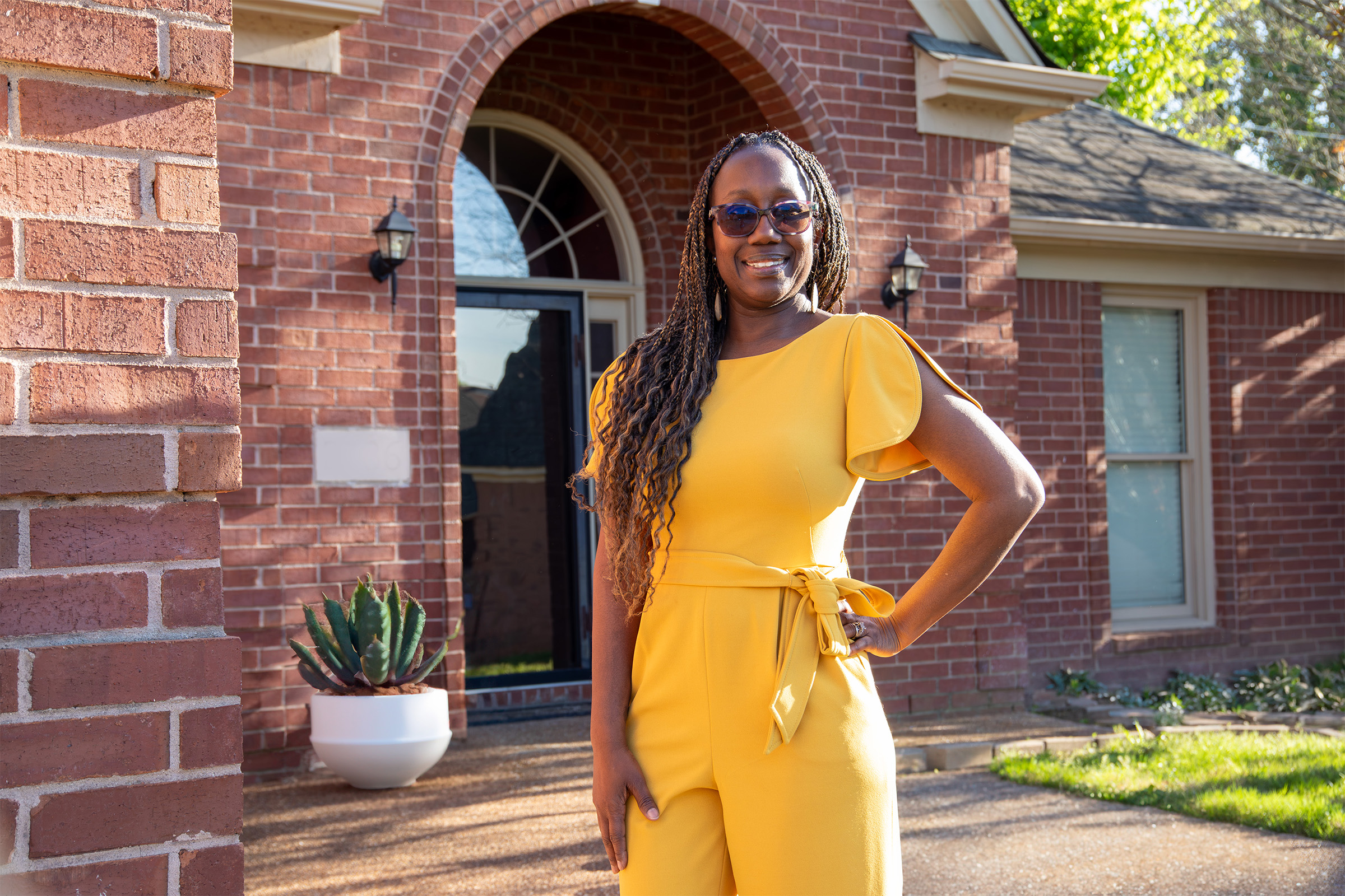 A woman wearing a yellow jumpsuit and sunglasses stands smiling with one hand on her hip in front of a red brick house with an arched doorway and a potted plant.