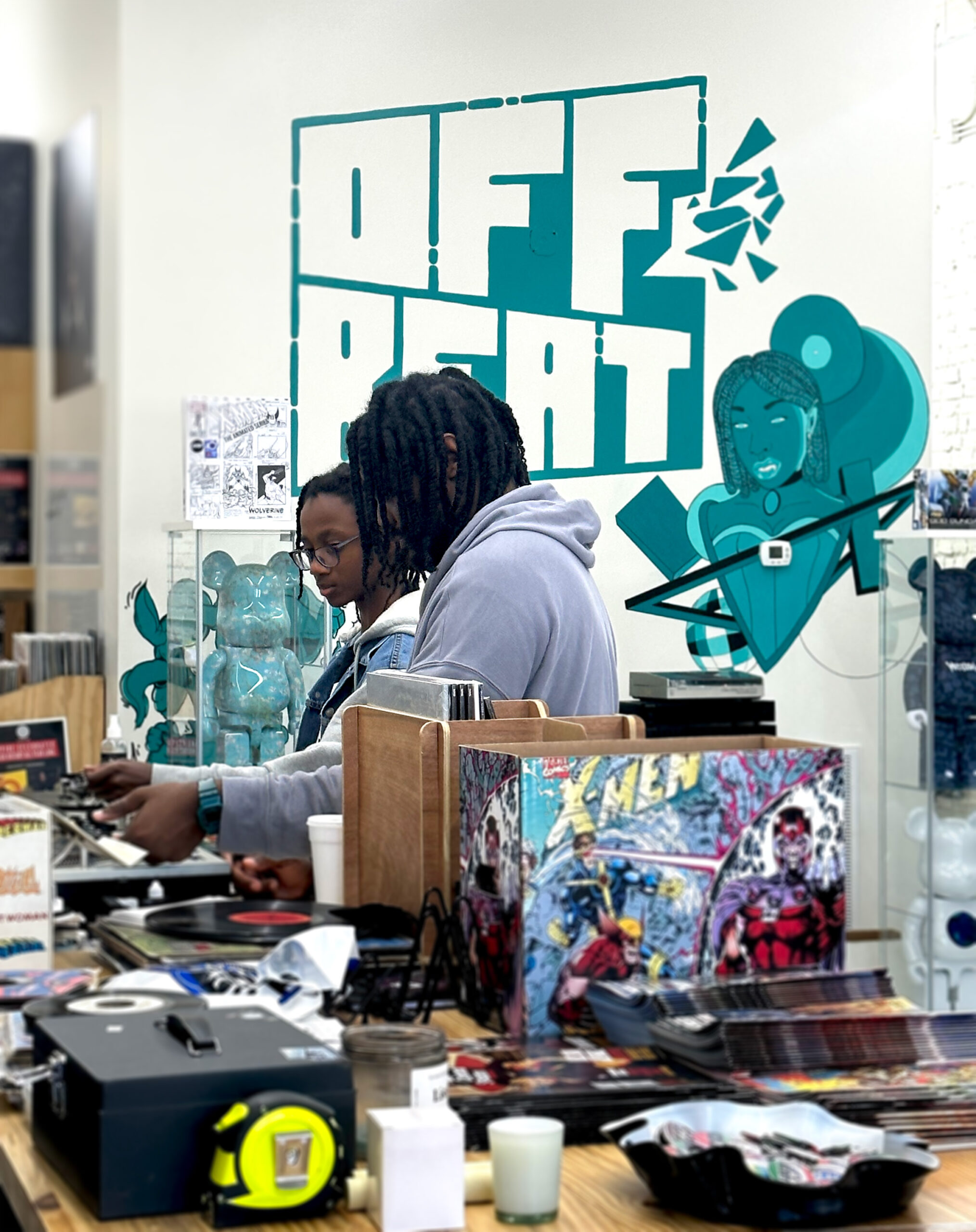 Two people stand behind a counter filled with comic books and collectibles at a store. A teal mural reading 