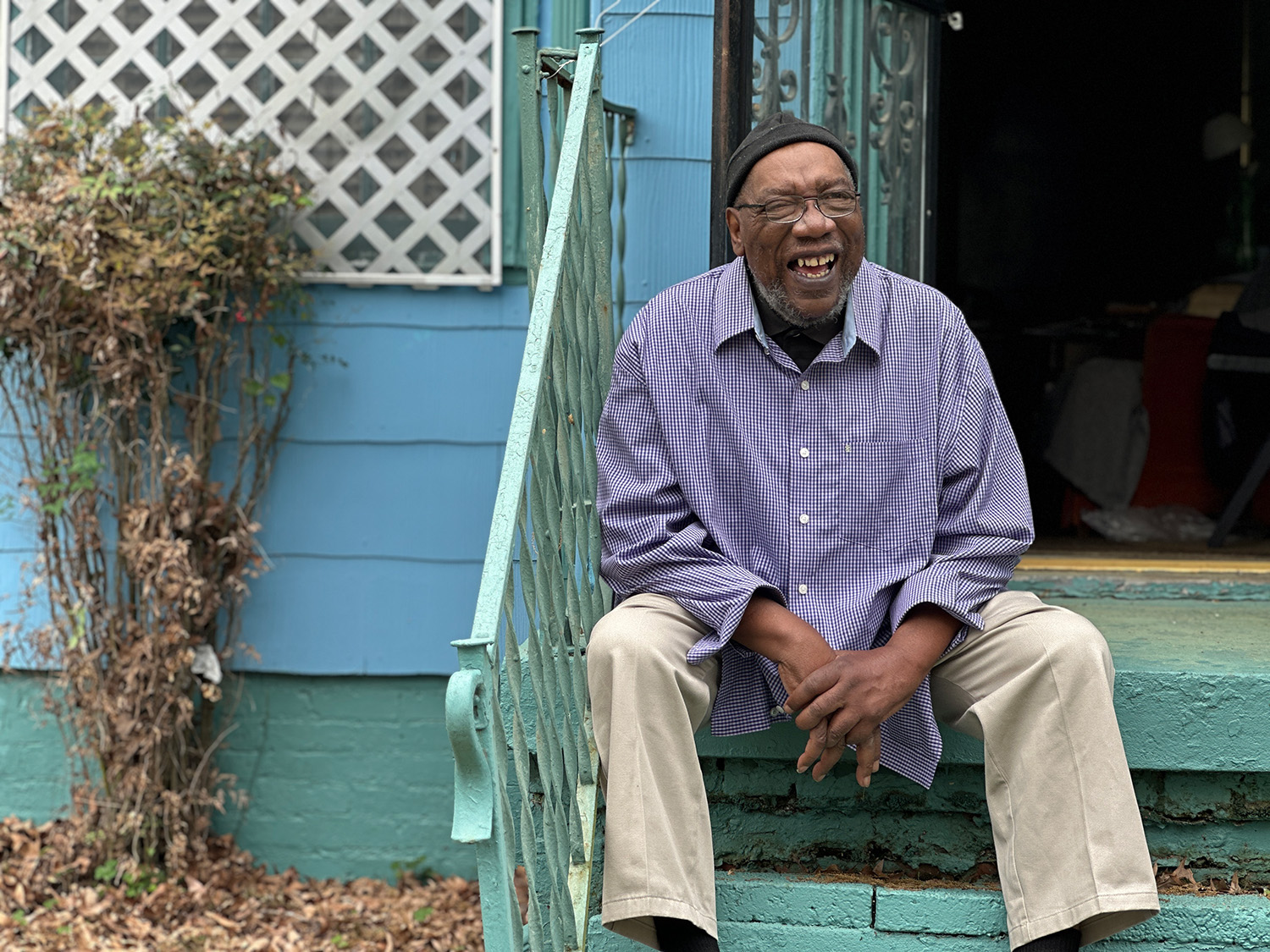 A man wearing glasses, a beanie, and a checkered shirt sits on the steps outside a blue house, smiling and laughing. Dry leaves and plants are visible to the left of the steps.