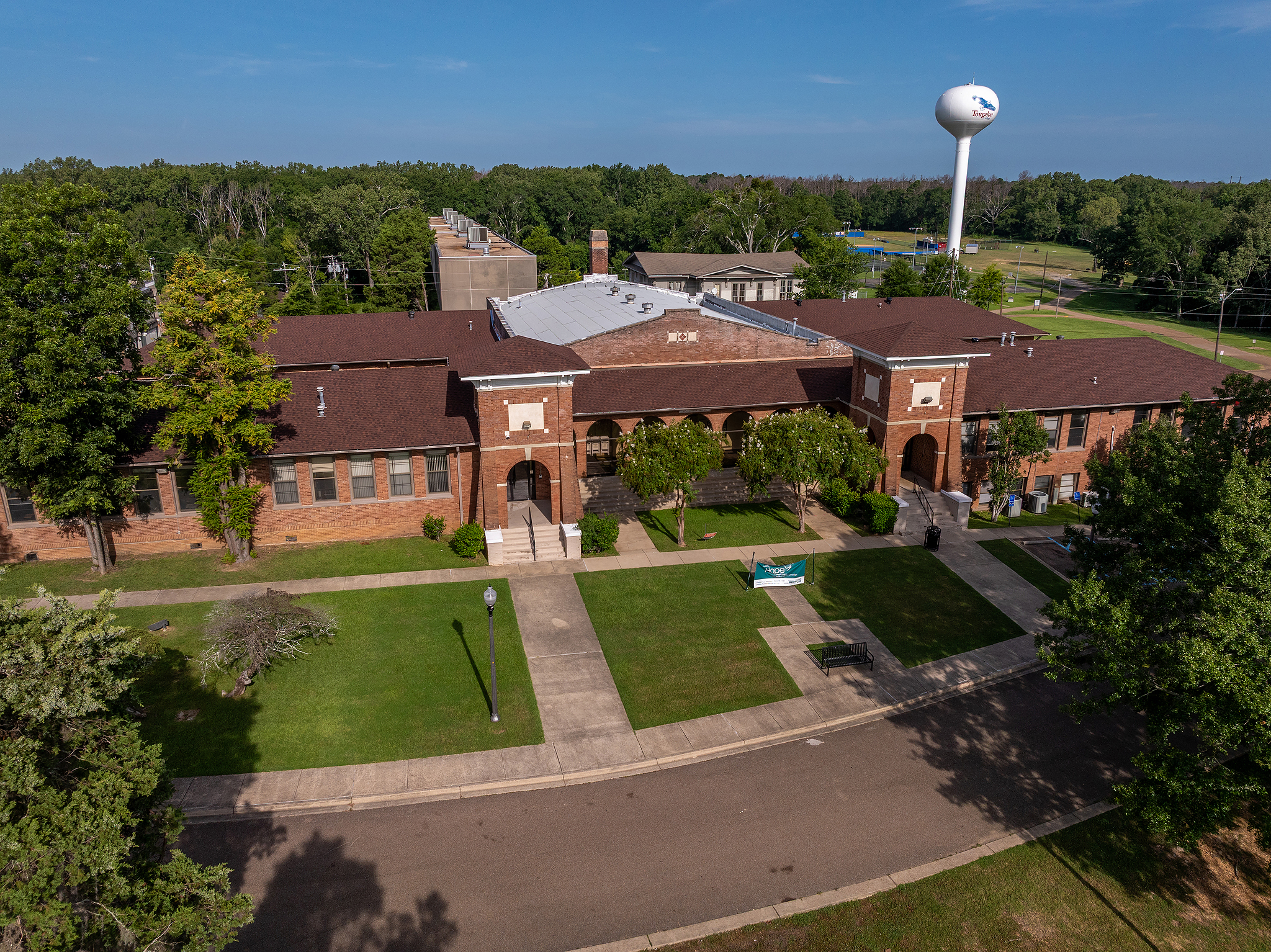 Aerial view of a large brick building with a central entrance, surrounded by trees and green lawns. A sidewalk curves in front, and a white water tower is visible in the background against a blue sky.