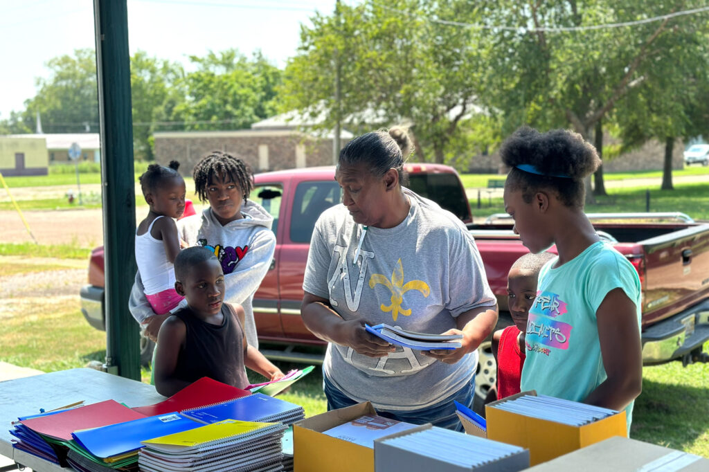 A woman hands out notebooks and folders to children at an outdoor table, with school supplies spread out in front, while a truck and trees are visible in the background.