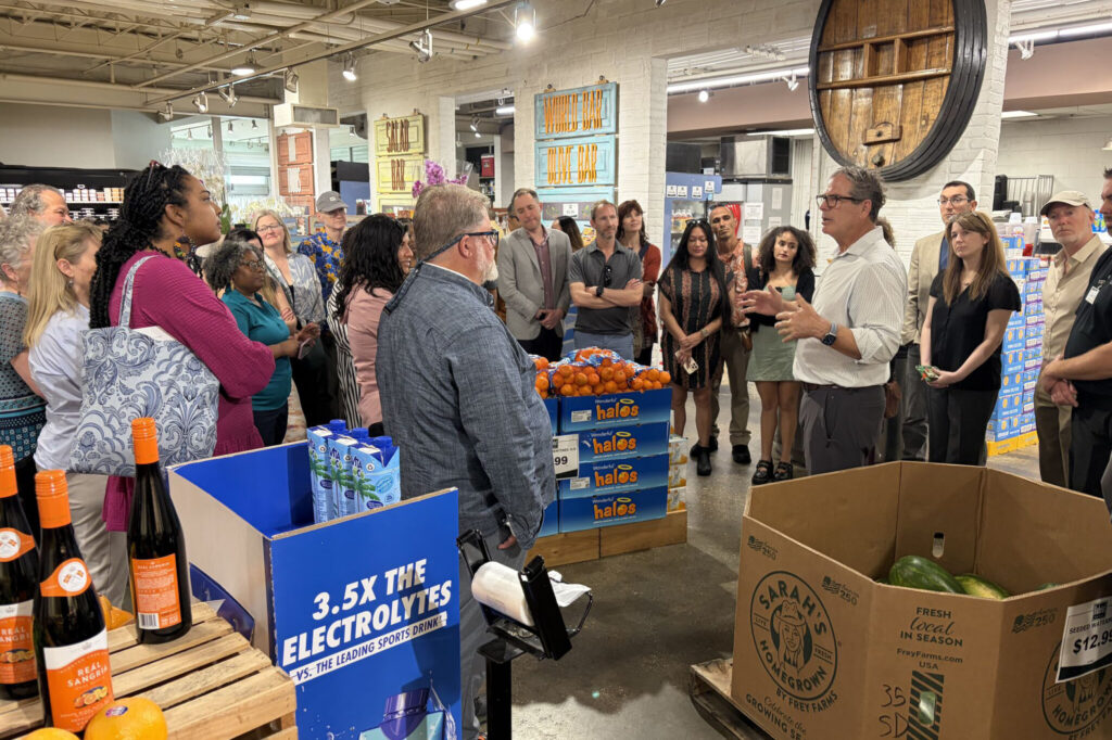 A group of people stands in a grocery store, listening to a man speak near large cardboard boxes of oranges and bottled drinks. The setting appears casual and informational, with products and displays visible in the background.