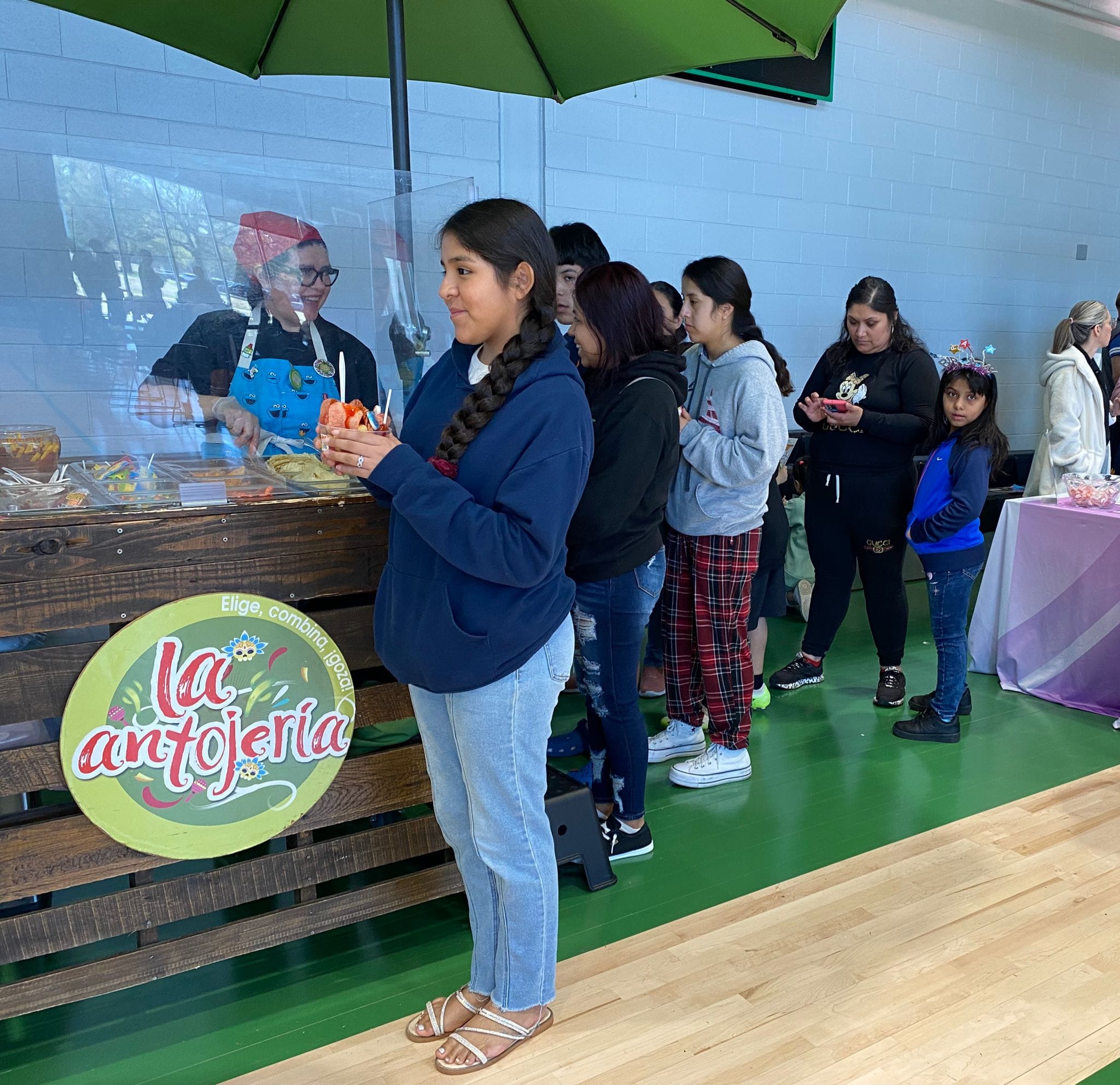 A group of people, including children, stand in line at a food stand labeled 