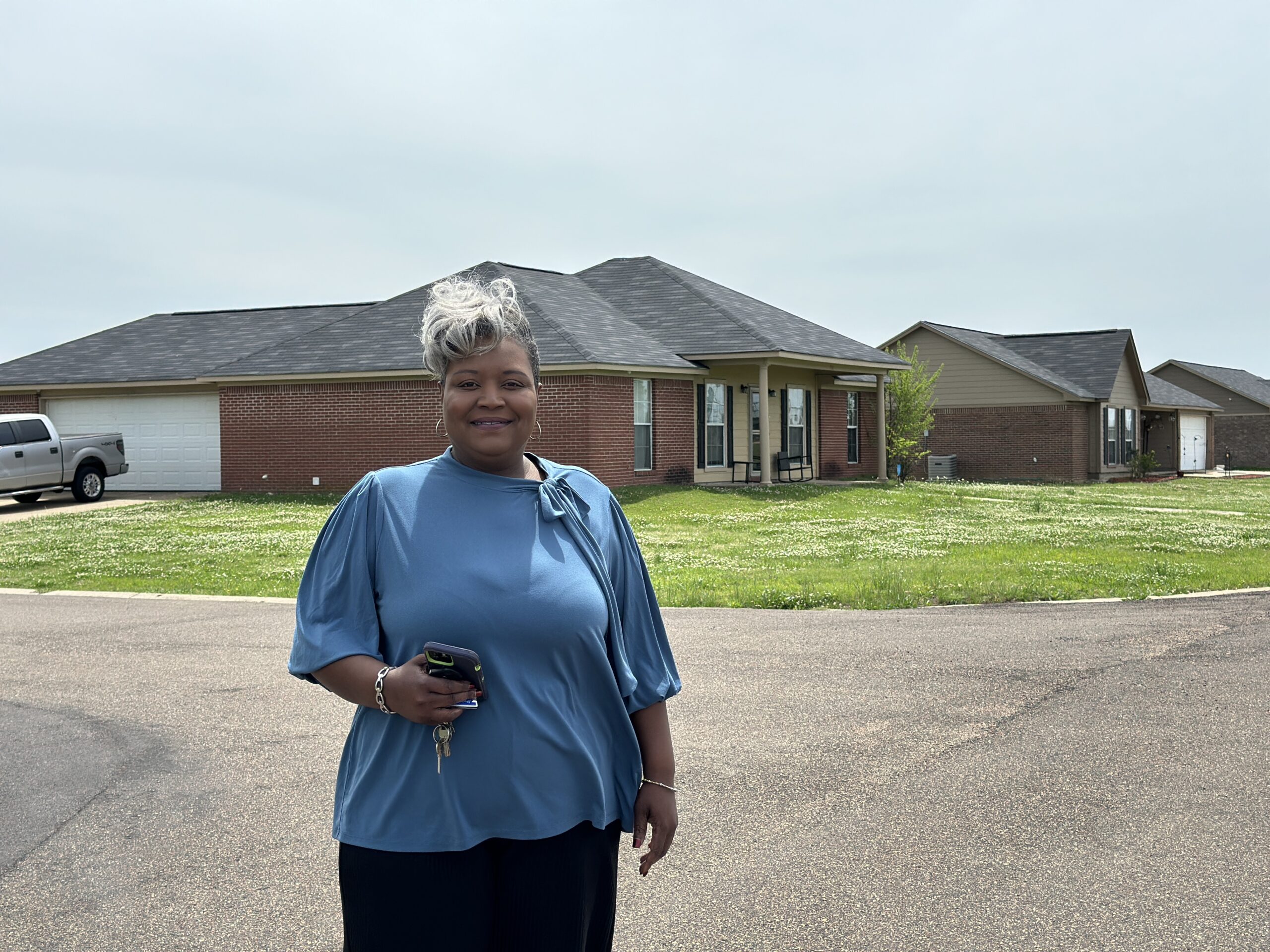 A woman with short gray hair, wearing a blue top, stands smiling on a suburban street holding keys and a phone. Behind her are single-story brick and tan houses, grass yards, and a cloudy sky.