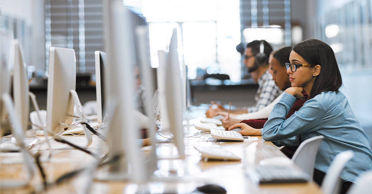 Several people are seated at a row of computers in a bright, modern workspace, working intently. The focus is on a woman wearing glasses and a blue shirt in the foreground.